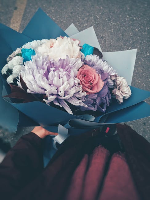 A vibrant floral bouquet arranged with large, fluffy white and purple chrysanthemums, complemented by soft pink roses and smaller blue flowers, wrapped in elegant blue and white paper with dark ribbon