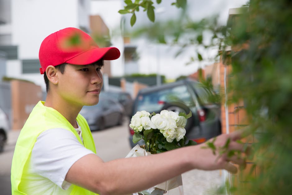 A young delivery person with light skin, wearing a red cap and a bright yellow high-visibility vest over a white t-shirt, is shown arranging a bouquet of fresh white flowers, possibly roses or peonies